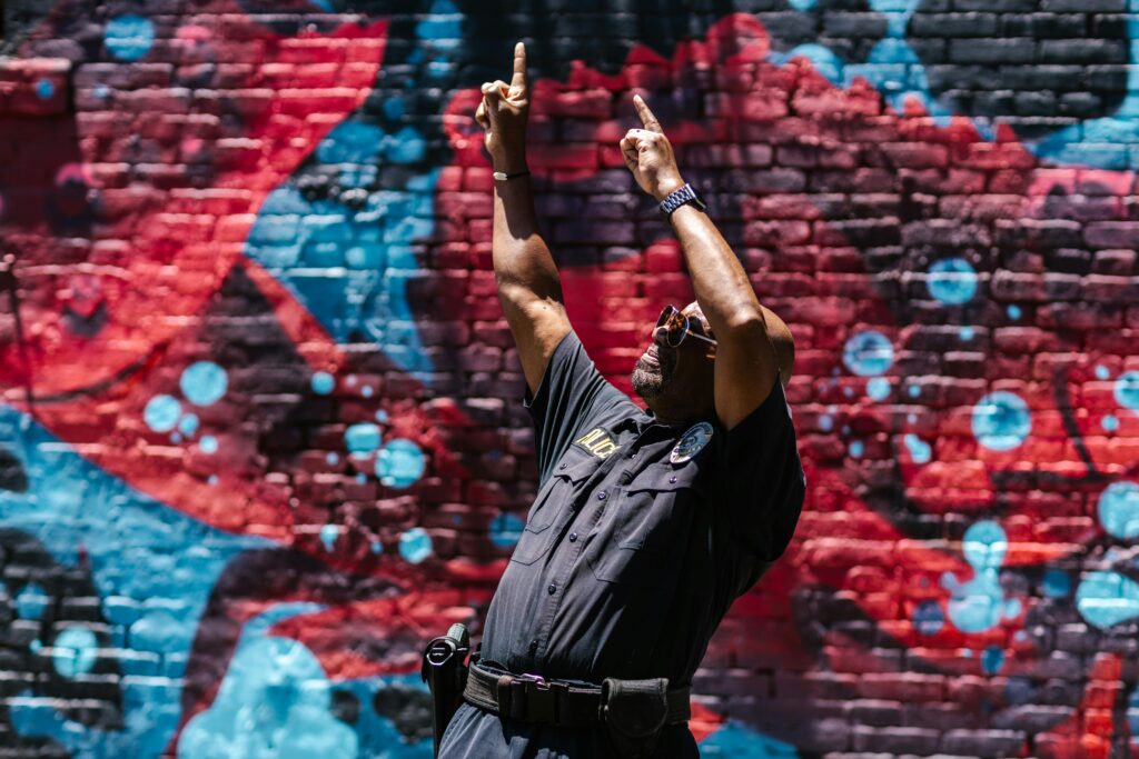 An African American man in police uniform dances in front of a colorful graffiti wall, showcasing joyful motion.